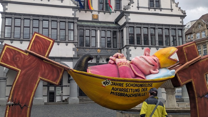 Ein Jacques Tilly Wagen sorgt für Aufsehen vorm Paderborner Rathaus. Der Wagen zeigt einen Bischof der in einer Hängematte ausruht. Aufschrift: 16 Jahre schonungslose Aufarbeitung der Missbrauchsfälle.