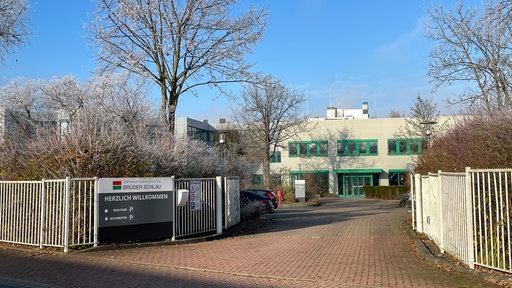 Ein graues Firmengebäude mit grünen Fenstern, die Bäume sind winterlich vereist, Firmenschild "Brüder Schlau - Herzlich willkommen"