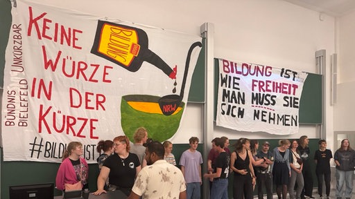 Rund 30 Studierende stehen in einem Hörsaal vor grünene Tafeln, darüber hängen Transparente