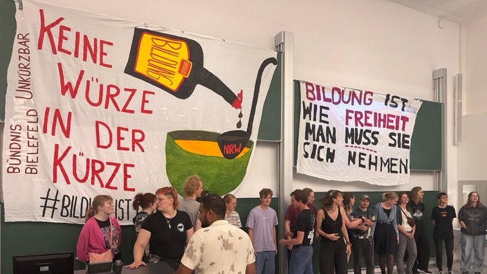 Rund 30 Studierende stehen in einem Hörsaal vor grünene Tafeln, darüber hängen Transparente