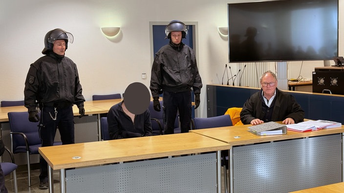 Der Angeklagte sitzt auf einem Stuhl. Neben ihm sein Anwalt. Hinter den beiden stehen zwei Wachbeamte.