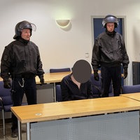 Der Angeklagte sitzt auf einem Stuhl. Neben ihm sein Anwalt. Hinter den beiden stehen zwei Wachbeamte.