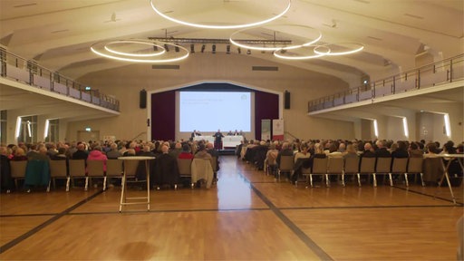 mehrere Menschen sitzen in einem Saal auf Stühlen und gucken auf eine Bühne