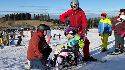 Eine Person sitzt auf einem Schlittena auf einer Skipiste. Mehrere Menschen stehen um sie herum 