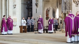 Geistliche in lilafarbenem Ornat stehen im Altarraum einer grauen Steinkirche, neben ihnen stehen Soldaten in grauer Uniform.