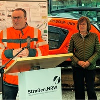 Verkehrsminister Oliver Krischer steht an einem Rednerpult. Hinter ihm sind Pläne zu sehen. Neben ihm steht eine Frau.