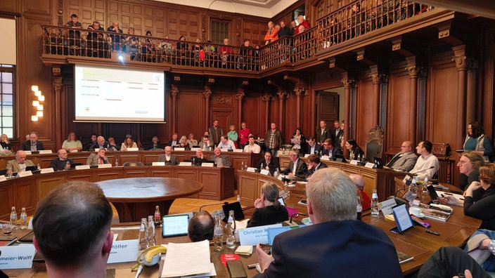 Zu sehen sind Politikerinnen und Politiker in einem Saal im Rathaus. Die Sitzbänke sind kreisförmig in mehreren Reihen angeordnet. Auf einem Balkon über den Politikerinnen und Politikern befinden sich viele Besucher. 