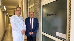 Zwei Männer stehen vor einer Tür auf der "Prosektur" steht. Einer von ihnen trägt einen Arztkittel, der andere einen Anzug 