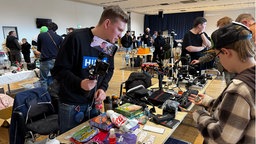 Eine Person steht an einem Tisch, auf dem unterschiedliche Dinge liegen, und filmt sich dabei mit selbst