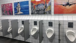 Über einer Reihe Pissoirs hängen verschiedene gerahmte Gemälde