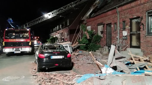 Vor einem beschädigtem Haus steht ein Pkw und ein Auto der mit Drehkran