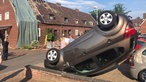 In einer Auffahrt liegt ein auf sein Dacht gekippts Auto, im Hintergrund stehen Leute auf der Straße