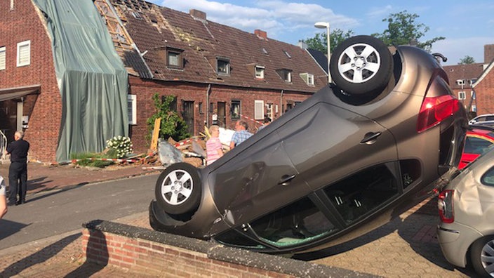In einer Auffahrt liegt ein auf sein Dacht gekippts Auto, im Hintergrund stehen Leute auf der Straße