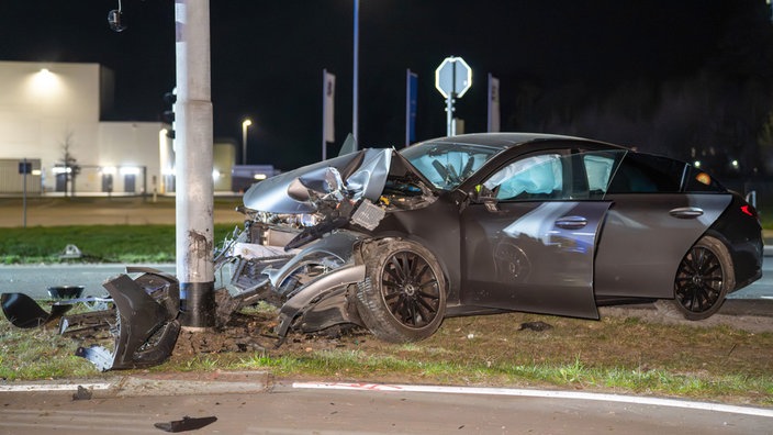 Auf einer Straße steht ein graues Unfallauto mit zerstörtem Motorraum, nachdem es gegen einen Ampelmast gefahren ist. Links vor dem Fahrzeug liegen Trümmer.