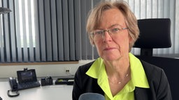 Eine ältere Frau mit randloser Brille, gelber Bluse und dunklem Blazer am Schreibtisch