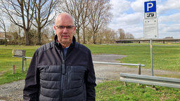 Ein Mann mit Brille, der vor einer Wiese steht. Auf einem Parkschild neben ihm steht die Aufschrift "Wohnmobilstellplatz".