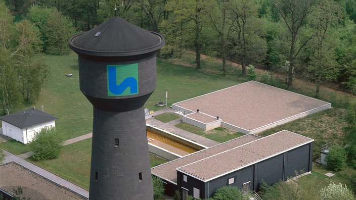 Ein Wasserturm steht im Vordergrund, im Hintergrund Gebäude und eine grüne Fläche