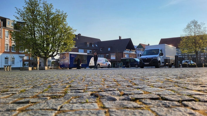Ein leerer Platz mit Pflastersteinen, auf dem hinten vereinzelt Transporter und Marktstände stehen.