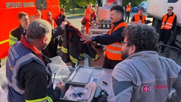 Eine Schutzjacke wird übergeben, im Hintergrund ein Fahrzeug der Feuerwehr Bonn.