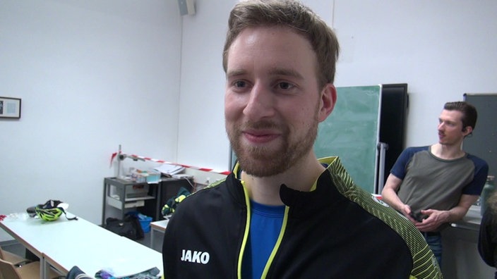Ein junger Mann mit Bart und Sportjacke steht in einem Raum mit einer Tafel an der Wand und lächelt