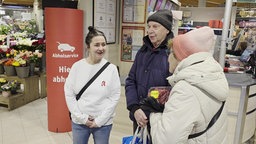 Eine Frau in einem Apothekenpulli unterhält mit zwei älteren Kunden in einem Supermarkt
