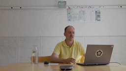 Ein Mann mit schütterem Haar und gelbem Poloshirt an einem Tisch mit Laptop und Wasserflasche