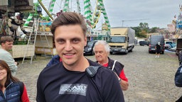 Ein junger Mann mit gegeelten Haaren, schwarzem Shirt und Funkgerät an der Schulter