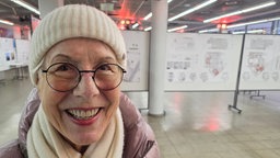 Eine ältere Frau mit runder Brille, weißer Mütze, weißem Schal und dicker Jacke vor einigen Stellwänden