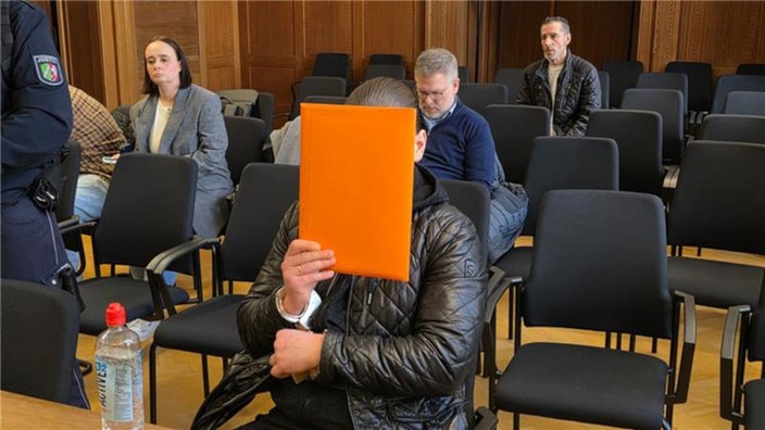 Der Angeklagte hält sich im Gerichtssaal einen orangen Aktenordner vor sein Gesicht.