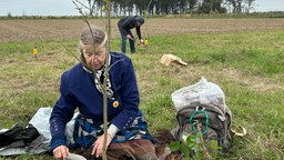 Eine Person pflanzt einen Baum ein. Im Hintergrund pflanzt eine weitere Person auch einen Baum ein