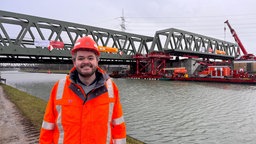 Ein Mann in Warnweste und mit Helm steht vor einem Kanal, im Hintergrund eine Brücke und Baukräne
