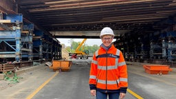 Eine Frau in orangener Warnjacke und mit Helm auf dem kopf steht auf einer gesperrten Autobahn, im Hintergrund ist eine Baustelle.
