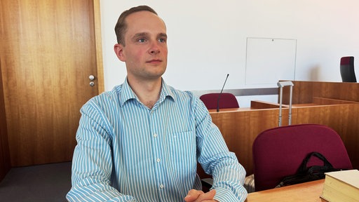 Der Bonner AfD-Politiker Gerald Christ, ein blondhaariger Mann mit gescheitelten Haaren im blau-weiß gestreiften Oberhemd, sitzt hinter einem Holztisch und schaut nach rechts oben.