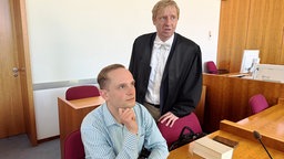 Der Bonner AfD-Politiker Gerald Christ, ein blondhaariger Mann mit gescheitelten Haaren im blau-weiß gestreiften Oberhemd, sitzt hinter einem Holztisch, rechts neben ihm steht sein Strafverteidiger in einer dunklen Robe.