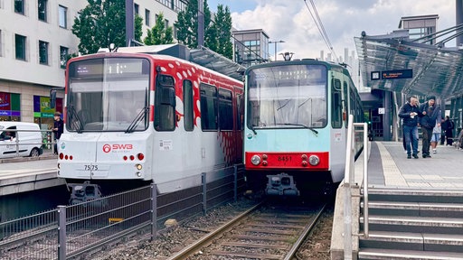 Zwei Stadtbahnen der Stadtwerke Bonn stehen auf den Gleisen der Haltestelle Stadthaus. Auf dem Bahnsteig laufen Menschen.
