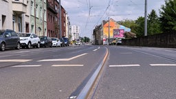 Auf einer zweispurigen Hauptstraße liegen Schienen der Bonner Stadtbahn. Links parken Autos hintereinander, rechts ist eine Backsteinmauer.