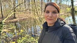 Eine Frau, Saskia Warner, steht in einem Wald vor einem Teich