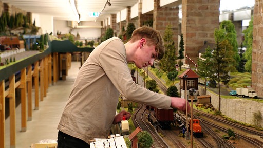 Ein junger Mann in beigem Langarmshirt greift zu einer Modelleisenbahn. Im Hintergrund ist eine weitläufige Modelleisenbahnstrecke aufgebaut.