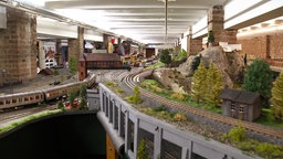 In einem großen Raum ist weitläufig eine Modelleisenbahnstrecke mit Miniaturbäumen, -felsen und -häusern ausgelegt.