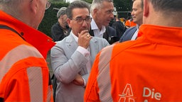 Der Bürgermeister von Bonn, Guido Déus, hält seine rechte Hand an sein Kinn. Déus trägt einen grauen Anzug und eine Brille. Vor ihm stehen zwei Mitarbeiter der Autobahn GmbH in orangenen Warnwesten, deren Gesicht man nicht sehen kann.