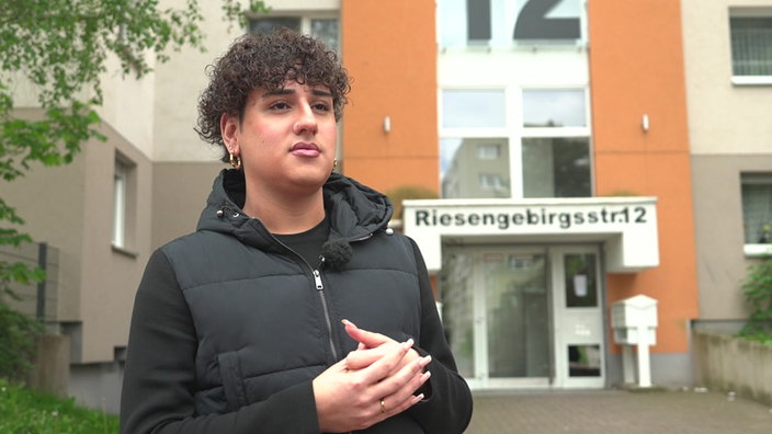 Ein Mann mit Locken steht vor einem orangenen Hochhaus. Er hat seine Hände gefalten, seine Nägel sind manikürt. Er trägt goldene Creolen. Seine Kleidung ist schwarz. 