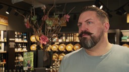 Mann mit dunklen Haaren und einem starken Schnauzbart steht in einem Geschäft. Hinter ihm sind Fässer und Flaschen mit Öl. 