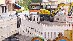 Weiße Leitbaken umzäunen eine Baustelle. Darin steht ein gelber Bagger. Auf der schmalen Fahrbahn am linken Bildrand stauen sich viele Fahrzeuge.