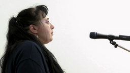 Eine Person mit schwarzen langen Haaren und Pony steht im Profil vor einem Mikrofon.
