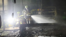 Zwei Feuerwehrmänner stehen in einem verkohlten Baseballkäfig und halten einen Schlauch aus dem Wasser kommt