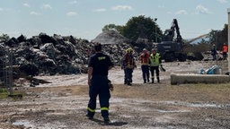 Ein Mitarbeiter des THW geht auf einige Kollegen der Feuerwehr zu, im Hintergrund werden Müllberge gelöscht