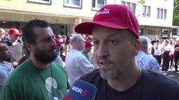 Ein Mann mit roter Kappe und Bart vor einer Menschenansammlung