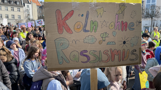 Im Vordergrund ein Plakat mit der Aufschrift "Köln ohne Rassismus". Im Hintergund befindet sich eine große Menschenmasse.