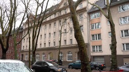 Eine breite Häuserfront von der Straße aus, davor Bäume