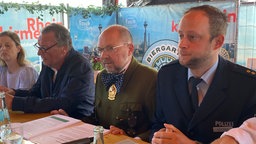 Auf dem Foto sind drei Männer, die an einem Tisch sitzen. Rechts einer im Anzug, links einer in Polizei-Uniform und in der Mitte Schützenchef Stieber in Schützen-Montur.